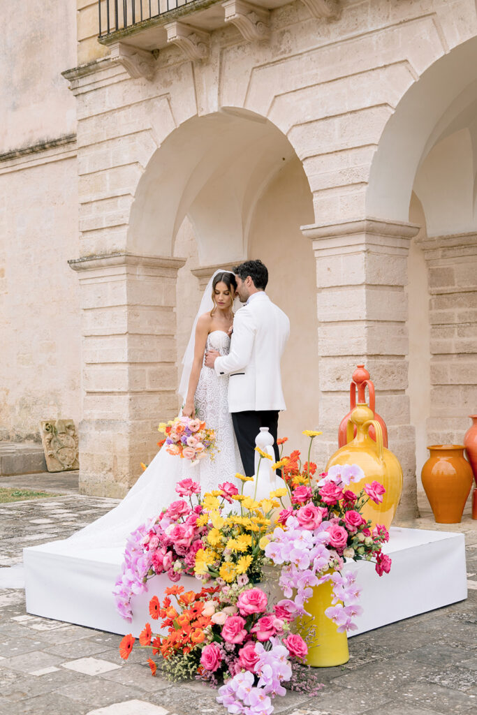 Fashion-forward bride and groom posing in front of vibrant coral and yellow ceremony florals.