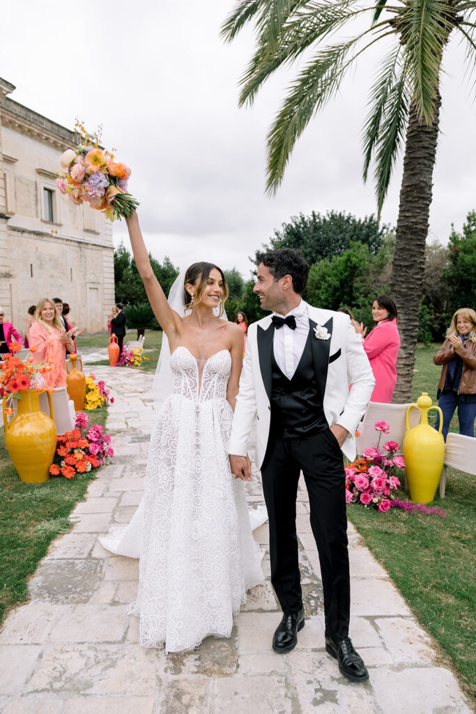 Bride raising colorful wedding bouquet in celebration alongside groom outdoors in Italy.