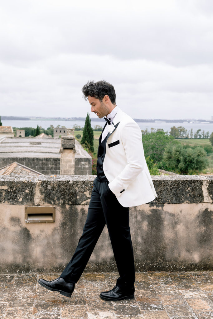 Fashion-forward groom in a sleek tuxedo posing against pale limestone walls for a Puglia destination wedding.