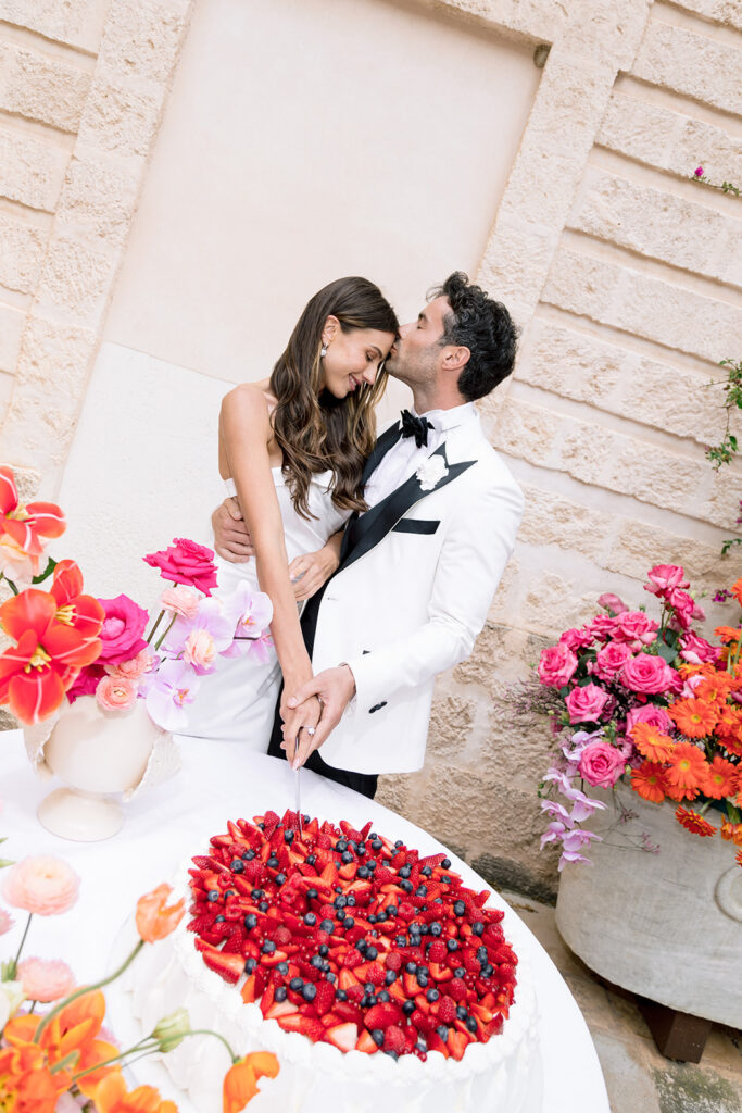 Groom kissing bride on forehead surrounded by bright pink and coral wedding flowers at Relais Histò in Puglia.